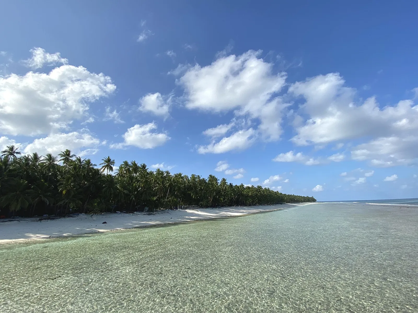 Clear shallow lagoon water over sand, with a palm-lined shore curving away under a cloudy blue sky