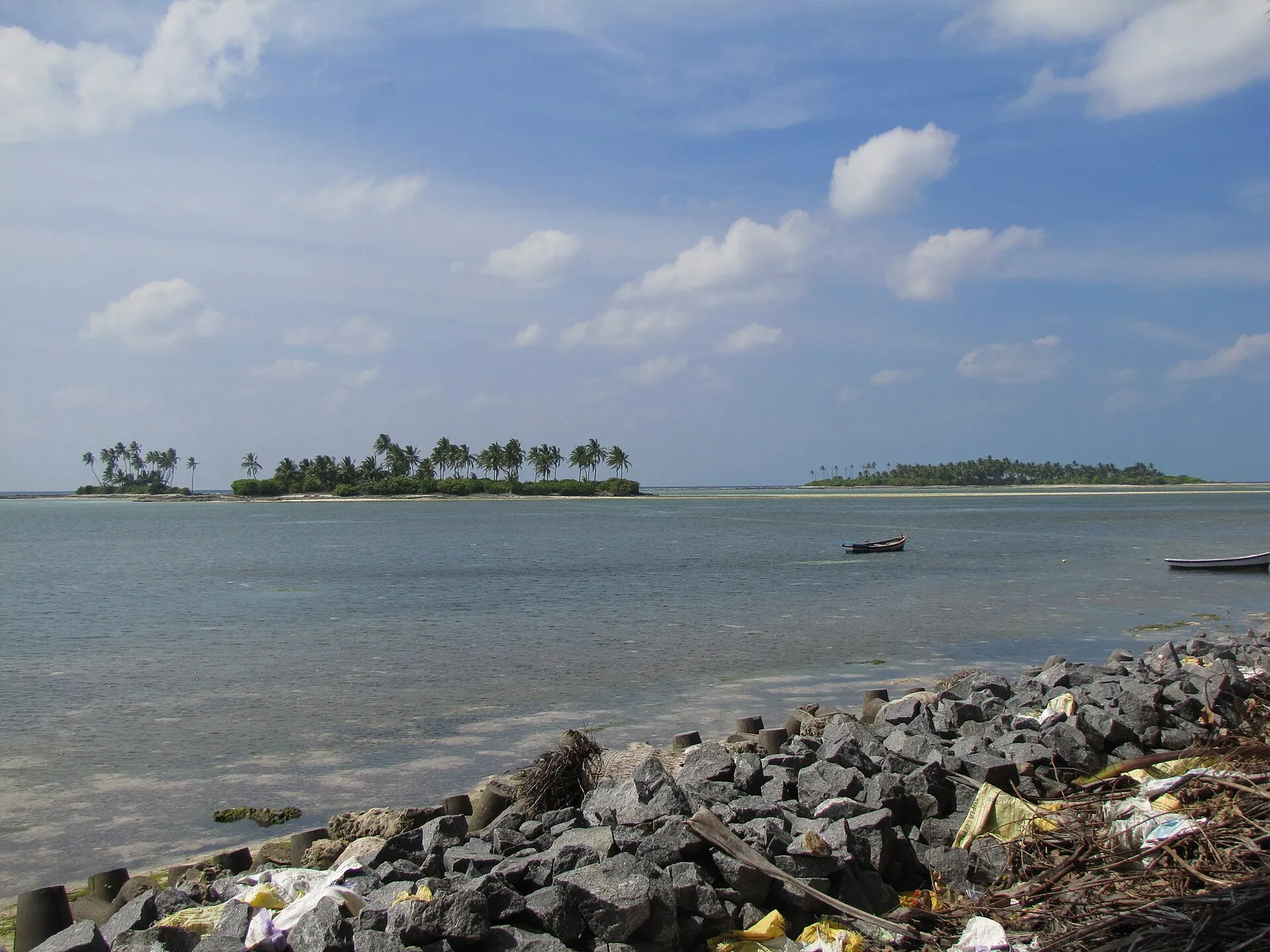 Small palm-covered islets and a moored boat in a calm lagoon, seen past a rocky shore