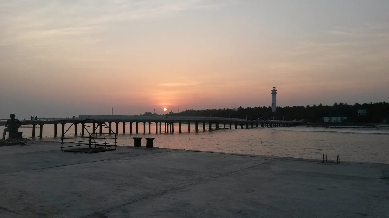 Sunset over a long jetty and low tree line, with a lighthouse tower on the skyline
